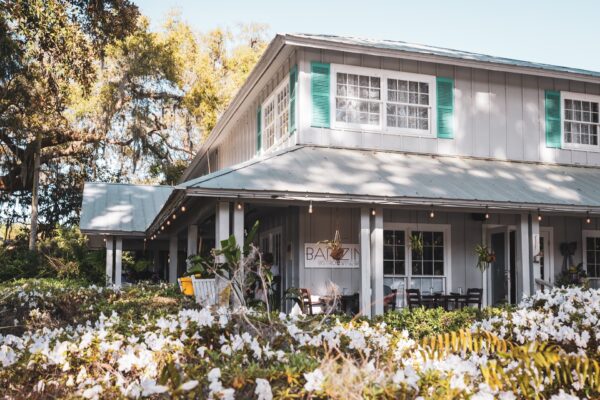 A charming gray building with teal shutters and a wraparound porch is surrounded by blooming white flowers and greenery. A sign reading Bar Zin hangs near the entrance under string lights.