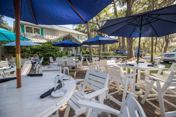 Outdoor dining area with white tables and chairs, shaded by blue umbrellas. Glasses and napkins are set on the tables. Greenery and a white house are visible in the background under a sunny sky.