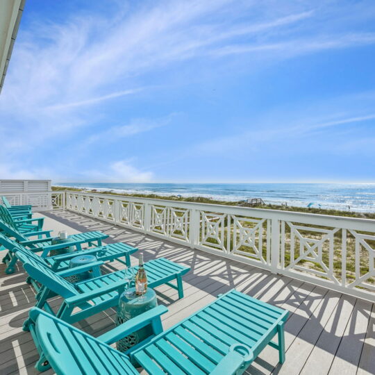 Turquoise lounge chairs sit on a spacious deck with a white railing, overlooking a sunny beach and ocean under a blue sky with wispy clouds.