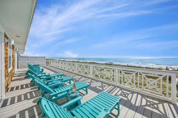 Turquoise lounge chairs sit on a spacious deck with a white railing, overlooking a sunny beach and ocean under a blue sky with wispy clouds.