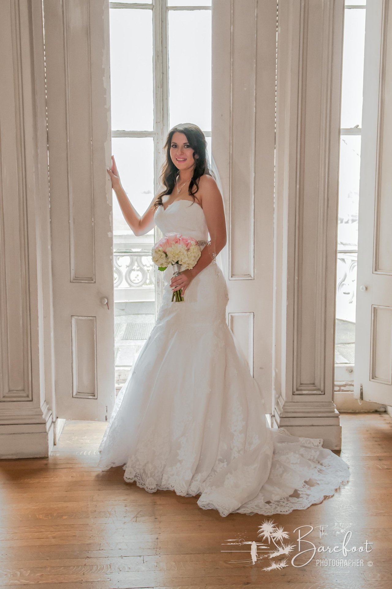 A bride in a white strapless wedding gown and veil stands by tall white French doors, holding a bouquet of pink and white flowers, with sunlight streaming in behind her.