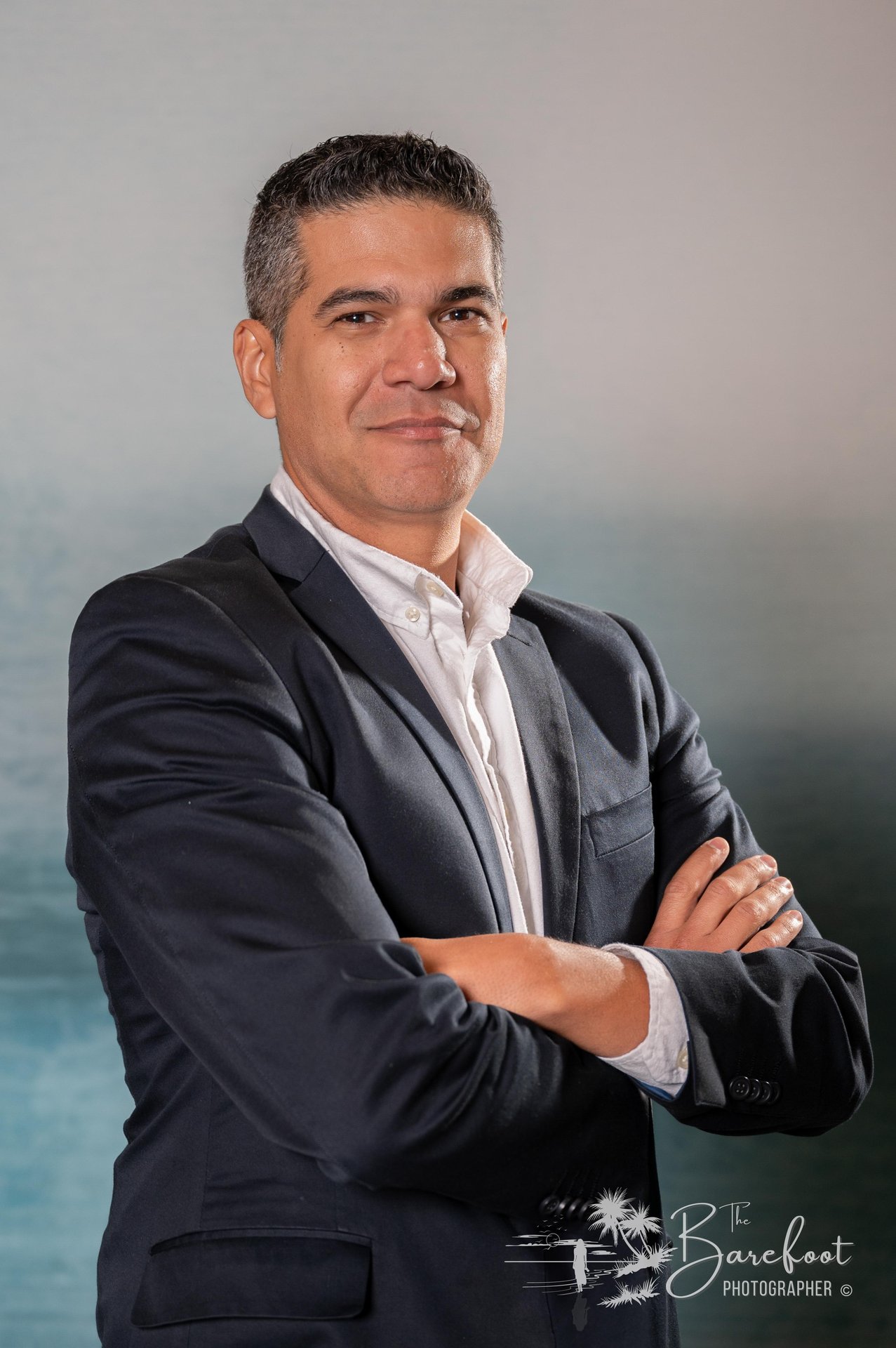 A man in a dark suit and white shirt stands with arms crossed, smiling slightly, in front of a soft blue and white background. A logo reading “The Barefoot Photographer” appears in the lower right corner.