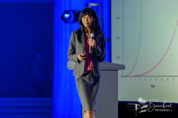 A woman in a gray suit stands on stage holding a microphone and a remote, with a screen behind her displaying a graph. Blue lighting illuminates the background.