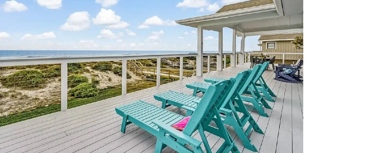A deck with turquoise chairs overlooks sand dunes and the ocean under a blue sky with scattered clouds.