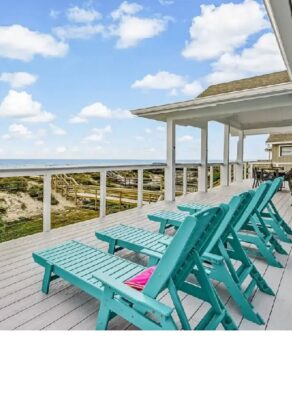 A deck with turquoise chairs overlooks sand dunes and the ocean under a blue sky with scattered clouds.