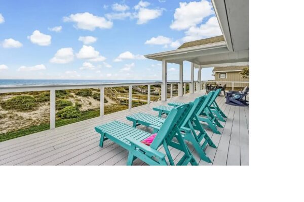 A deck with turquoise chairs overlooks sand dunes and the ocean under a blue sky with scattered clouds.