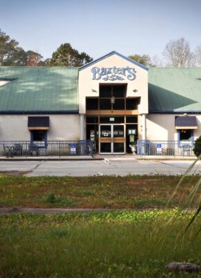 A restaurant named Baxters with a green roof and beige walls, seen from the parking lot, surrounded by trees and greenery on a sunny day.