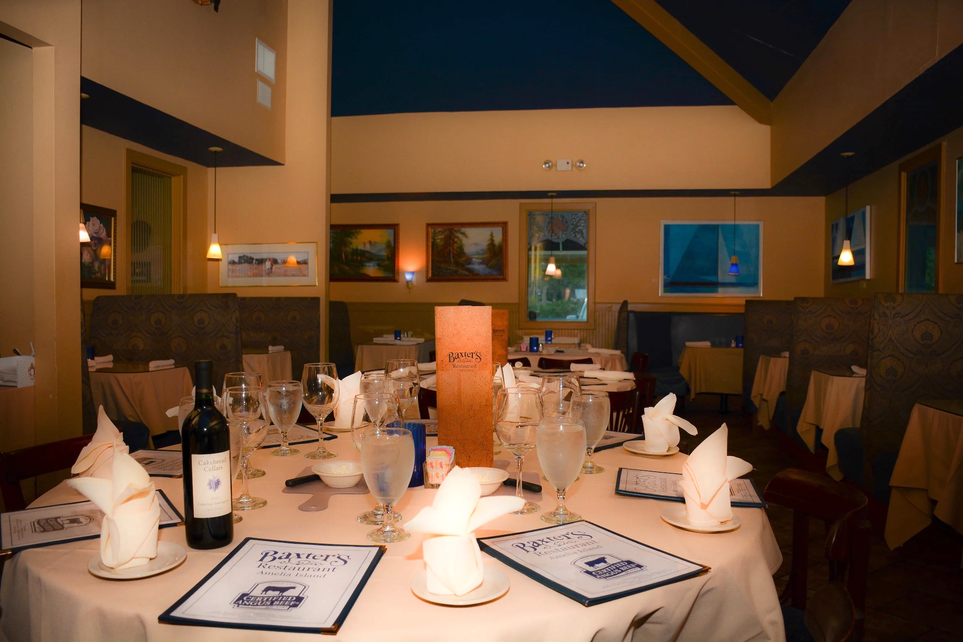 A set dining table at a restaurant with menus, wine, water glasses, napkins folded on plates, and a brown menu standing upright; booths and framed paintings are seen in the softly lit background.