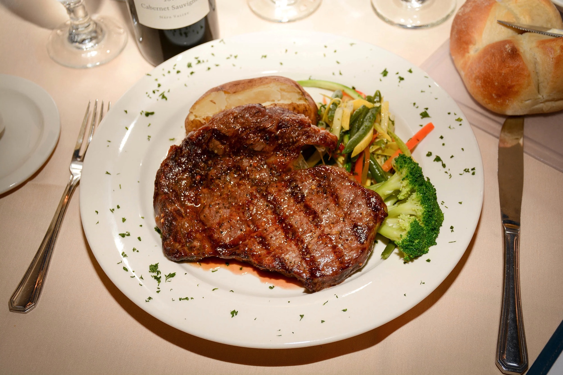 A grilled steak served on a white plate with a baked potato, steamed broccoli, and mixed vegetables, garnished with chopped herbs. A bread roll and wine glass are visible in the background.