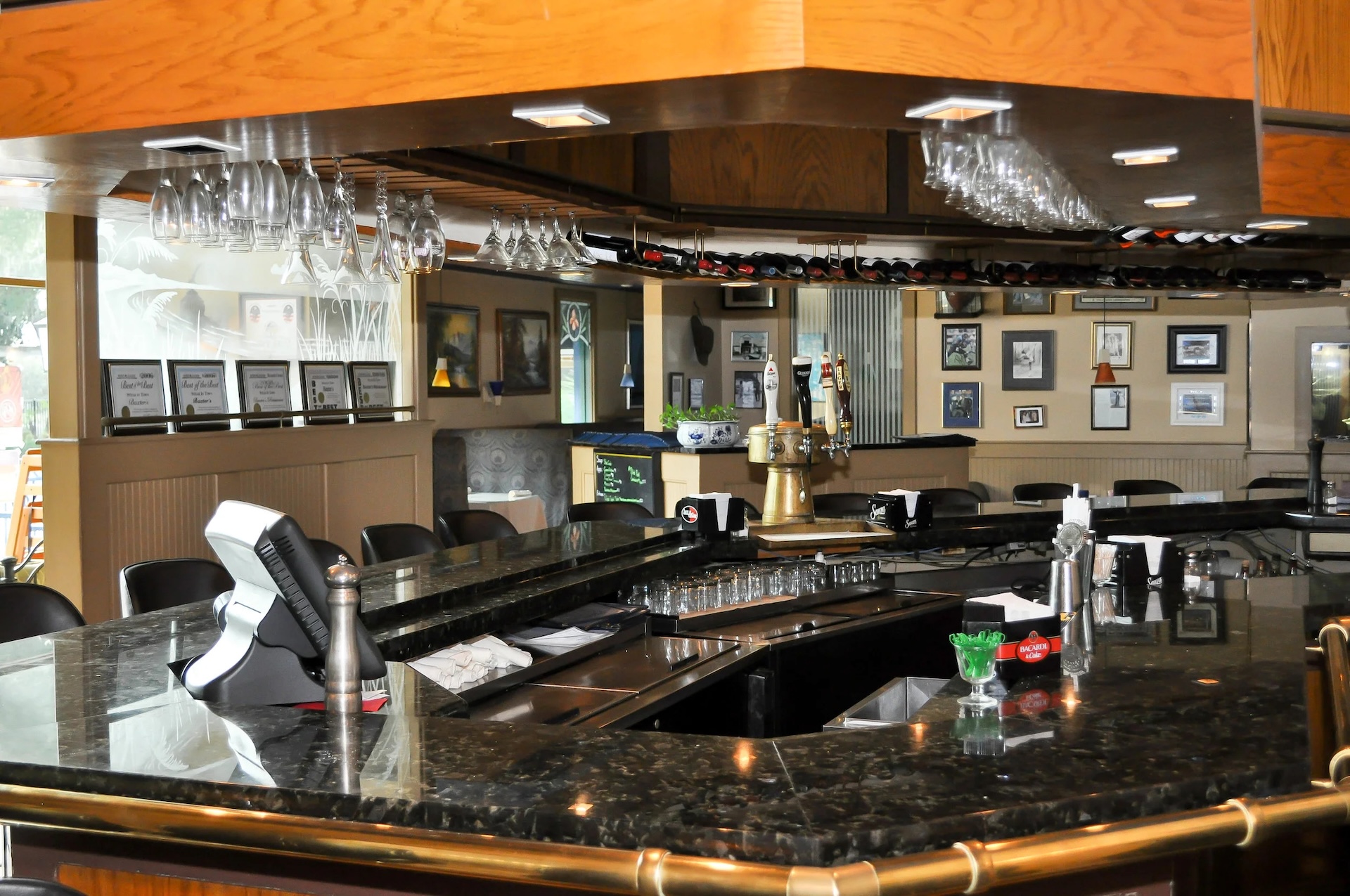 A polished black marble bar with barstools, hanging glasses, beer taps, and framed pictures on the walls in an empty, well-lit restaurant interior.