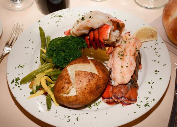 A plate with lobster tail, a baked potato, broccoli, green beans, and yellow beans, garnished with chopped parsley and a lemon wedge, served on a white tablecloth.