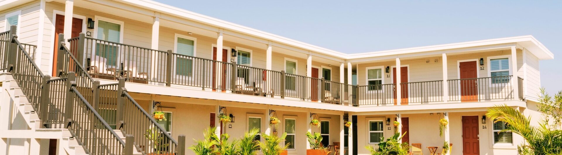 A two-story motel with exterior staircases, beige walls, and red doors. Flower planters and potted plants decorate the sunlit courtyard under a clear blue sky.