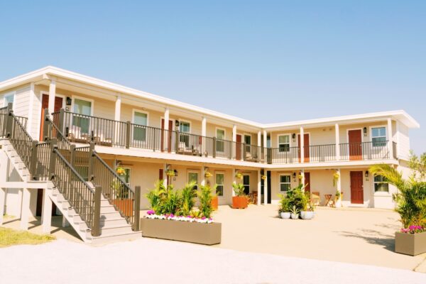 A two-story motel with exterior staircases, beige walls, and red doors. Flower planters and potted plants decorate the sunlit courtyard under a clear blue sky.