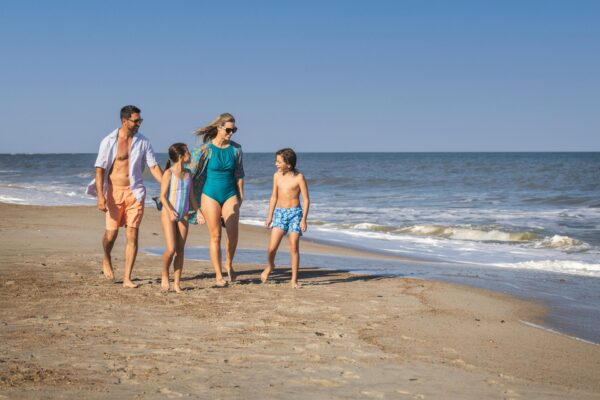 A family of four, two adults and two children, walks barefoot along a sandy beach near the ocean, smiling and enjoying a sunny day under a clear blue sky.