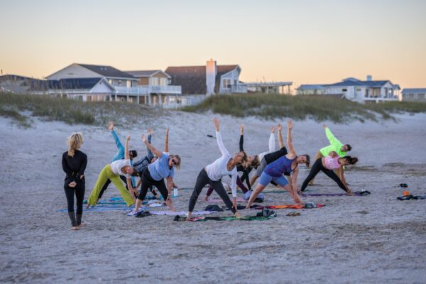 A group of people practice yoga on a sandy beach at sunset, stretching in a triangle pose while an instructor stands nearby. Houses and dune grass are visible in the background.