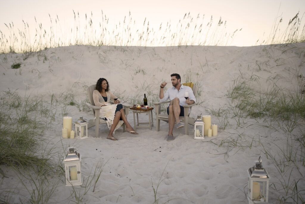 A man and woman sit in chairs on a sandy beach at sunset, sharing wine and snacks at a small table. Lit candles and lanterns surround them, creating a cozy and romantic atmosphere among sand dunes and grass.