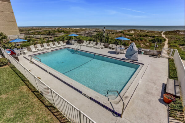 A fenced outdoor pool with lounge chairs and blue umbrellas overlooks grassy dunes and the ocean under a clear blue sky. A sandy path leads from the pool area toward the beach.