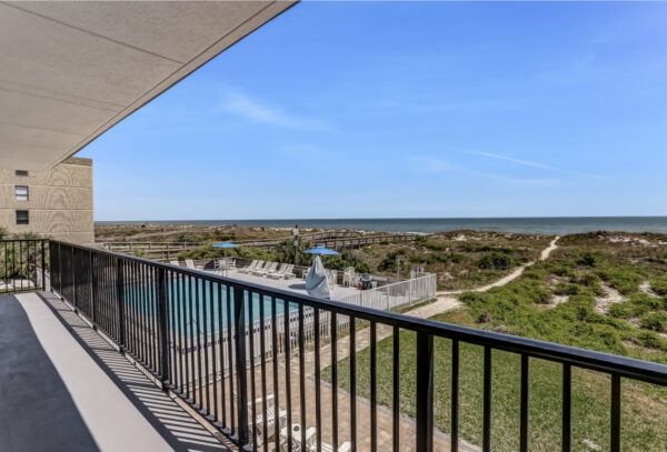 A balcony overlooks a pool area, sand dunes, and an ocean under a clear blue sky, with a path leading from the pool toward the beach.