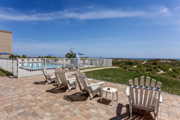 Several white Adirondack chairs sit on a brick patio facing grassy dunes and the ocean, with a fenced pool and blue sky in the background.