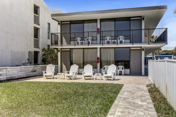 A modern two-story building with glass doors, balconies, several white chairs and loungers on a paved patio, and a small patch of green grass in front.