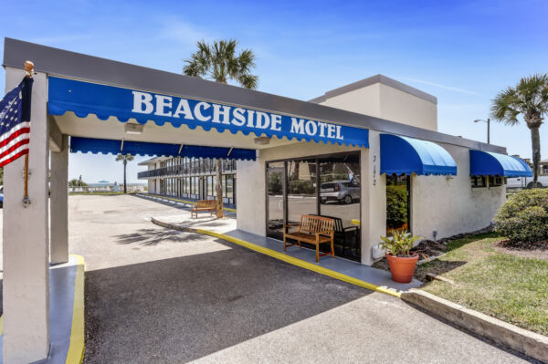 Beachside Motel exterior with blue awnings, glass doors, a wooden bench, potted plants, palm trees, and a partial view of the beach in the background under a clear sky.