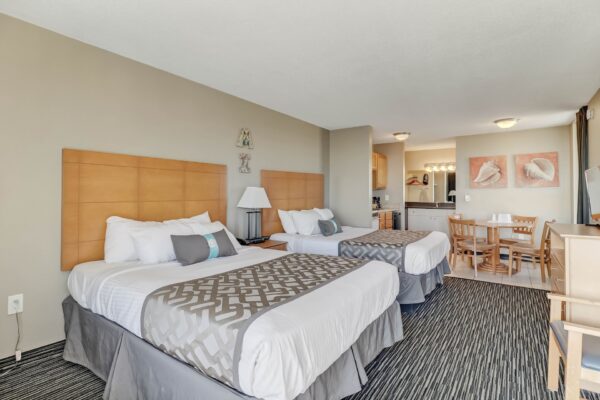 Hotel room with two double beds, light wood headboards, a small dining table with four chairs, kitchenette, desk, TV, and neutral-colored decor. Carpeting and modern lamps are visible, with artwork on the walls.