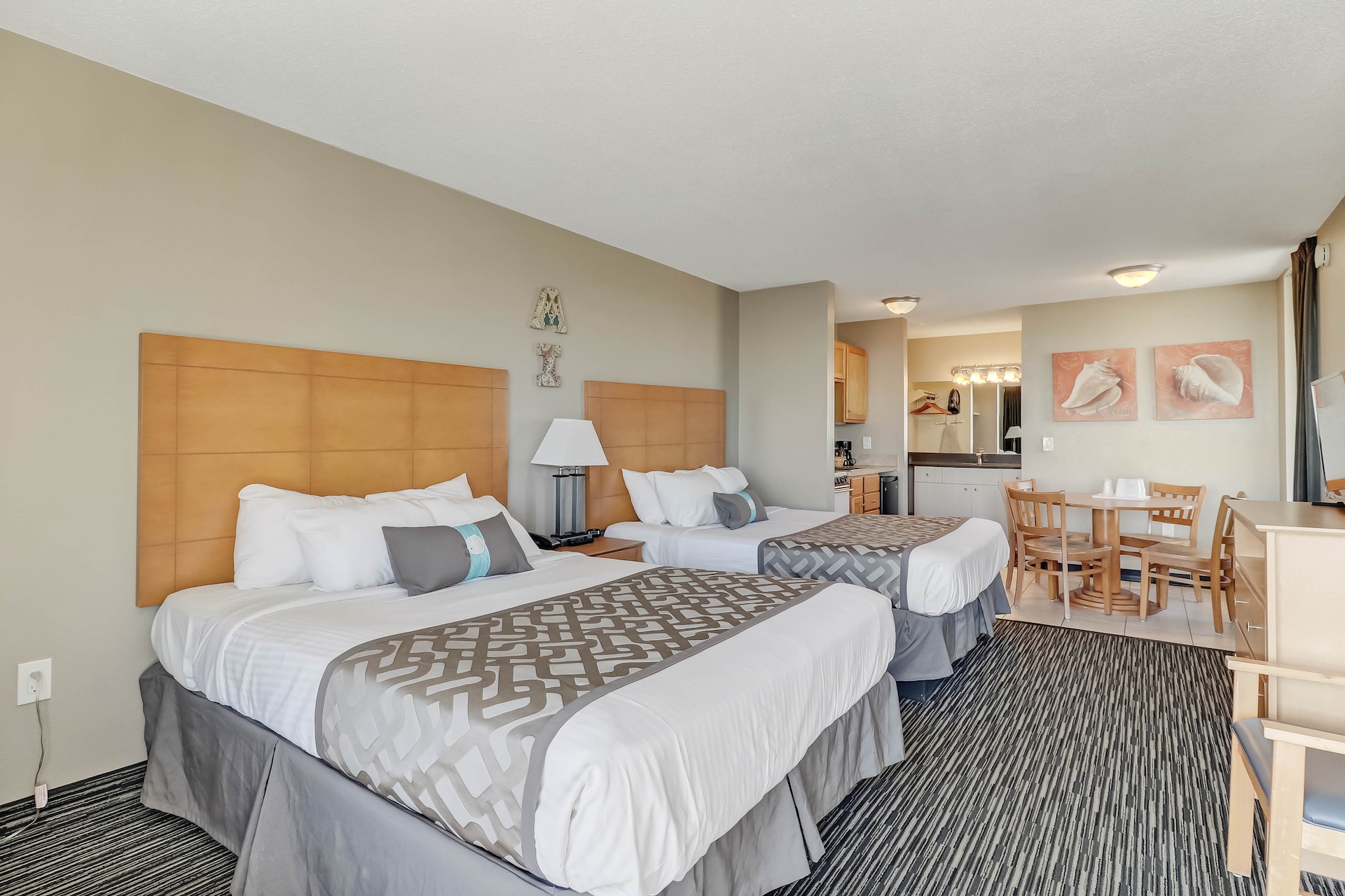 Hotel room with two double beds, light wood headboards, a small dining table with four chairs, kitchenette, desk, TV, and neutral-colored decor. Carpeting and modern lamps are visible, with artwork on the walls.