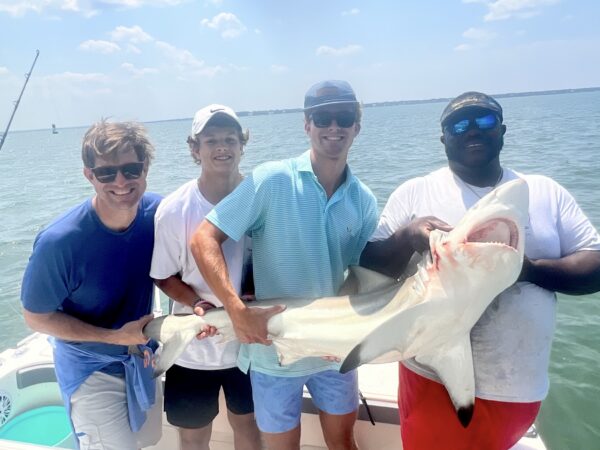 Four men on a boat smile at the camera while holding a large shark they caught, with the ocean and sky in the background on a sunny day.
