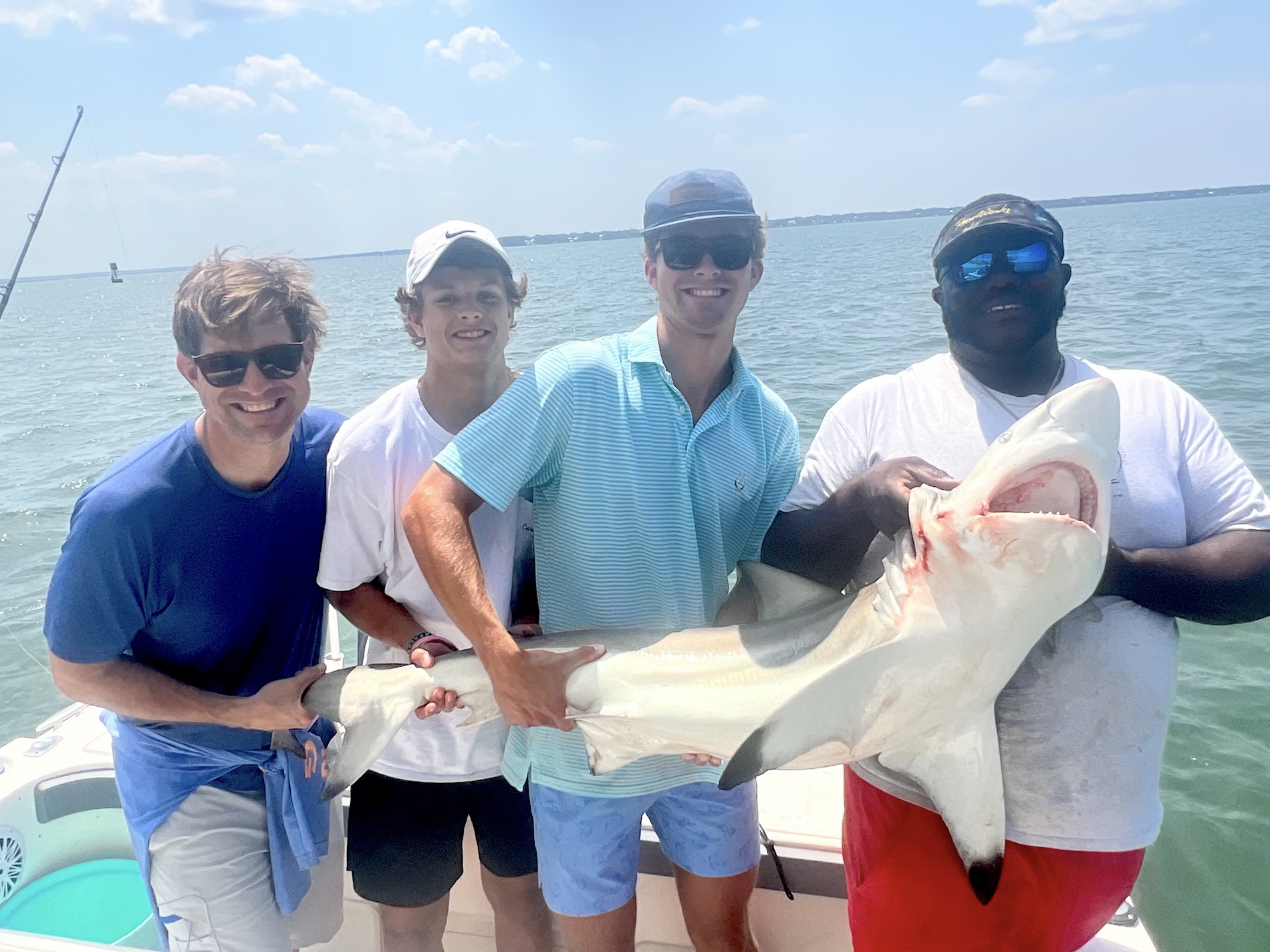 Four men on a boat smile at the camera while holding a large shark they caught, with the ocean and sky in the background on a sunny day.
