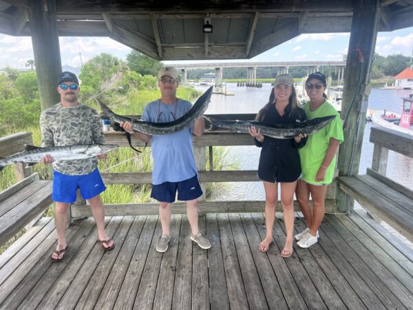 Four people stand on a wooden dock under a gazebo, with two men in the middle proudly holding large fish. Two women stand on either side smiling. A river and boats are visible in the background.