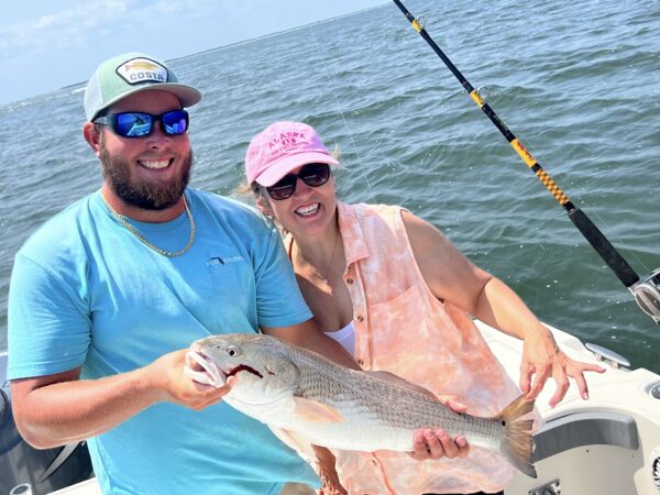 Two people on a boat smile while holding a large fish they caught. The man wears a blue shirt, cap, and sunglasses, and the woman wears a pink hat, sunglasses, and a sleeveless shirt. Water and a fishing rod are visible in the background.