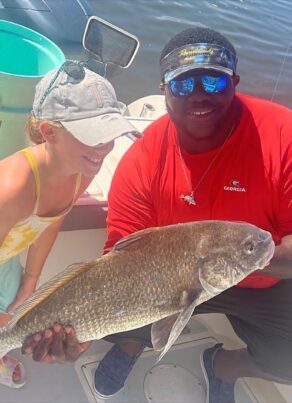 A young girl and a man on a boat smile while holding a large fish together. The man wears a red shirt and sunglasses; the girl wears a hat and colorful swimsuit. Fishing gear and a bucket are visible in the background.