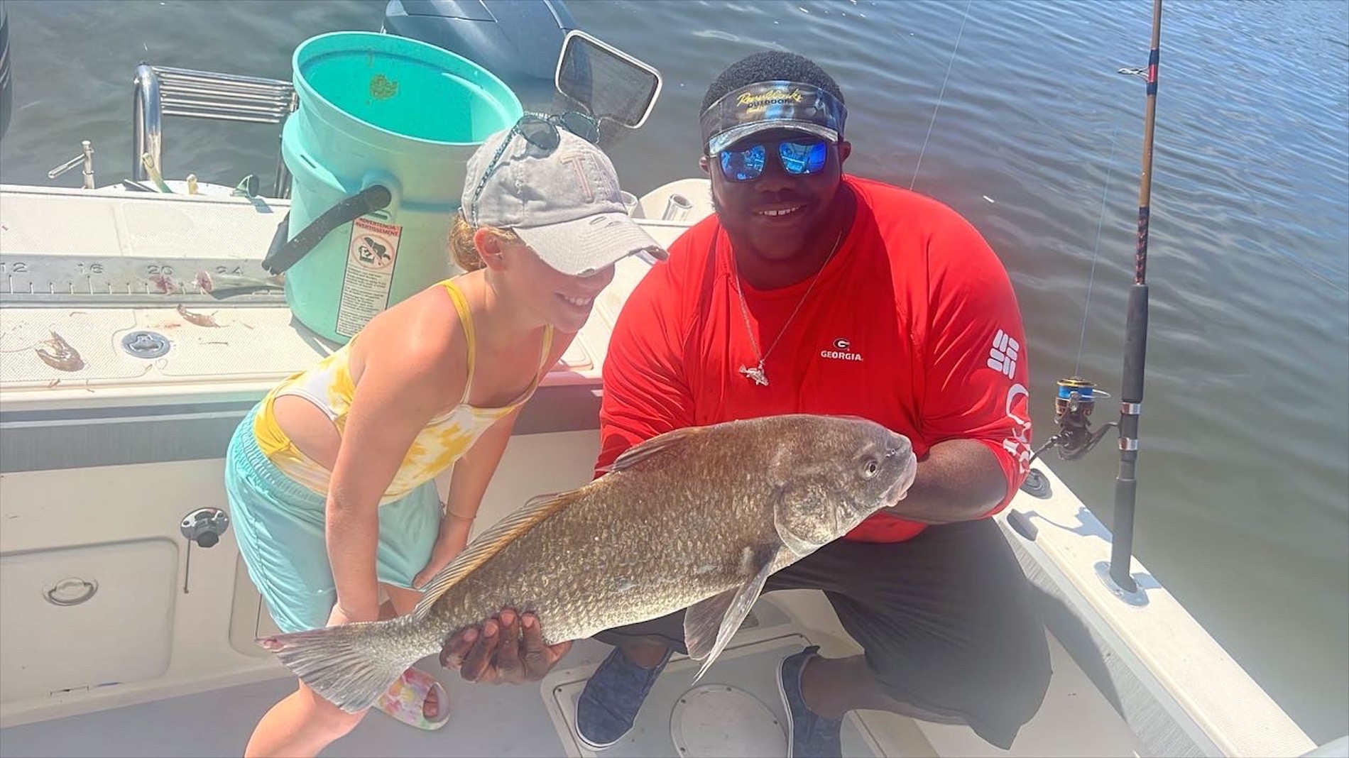 A young girl and a man on a boat smile while holding a large fish together. The man wears a red shirt and sunglasses; the girl wears a hat and colorful swimsuit. Fishing gear and a bucket are visible in the background.