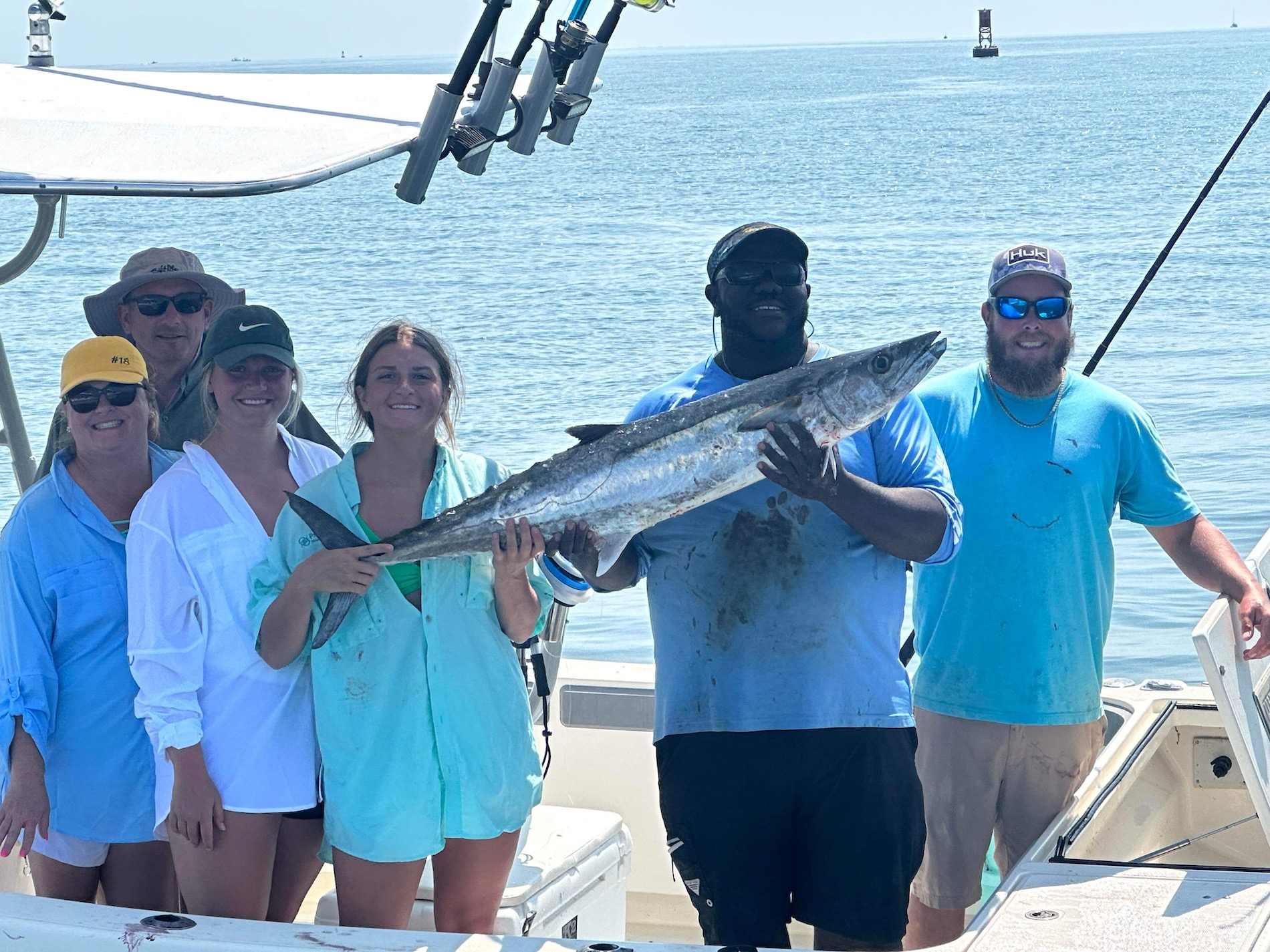 Five people on a boat smile at the camera as two of them hold a large fish. They are dressed in casual fishing attire, and the sea is calm in the background under a sunny sky.