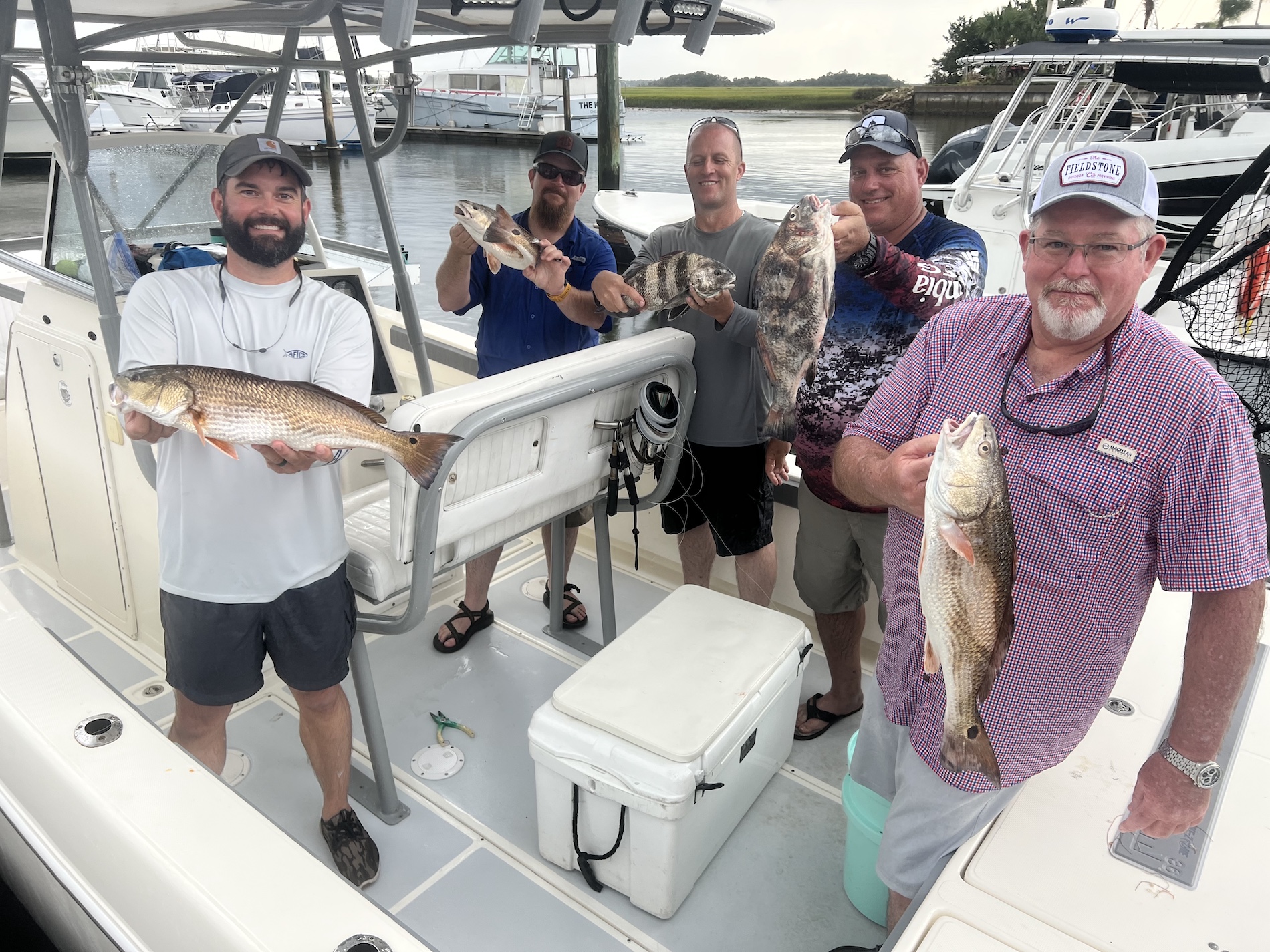 Five men on a boat docked at a marina proudly hold up their freshly caught fish, smiling at the camera. Various fishing equipment and other boats are visible in the background.