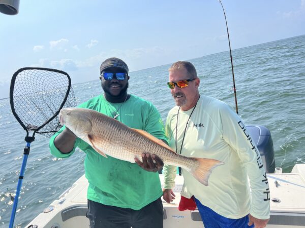 Two men on a boat at sea, one holding a large fish and the other holding a fishing net. Both are smiling and wearing sunglasses and long-sleeve shirts. A fishing rod is visible in the background.