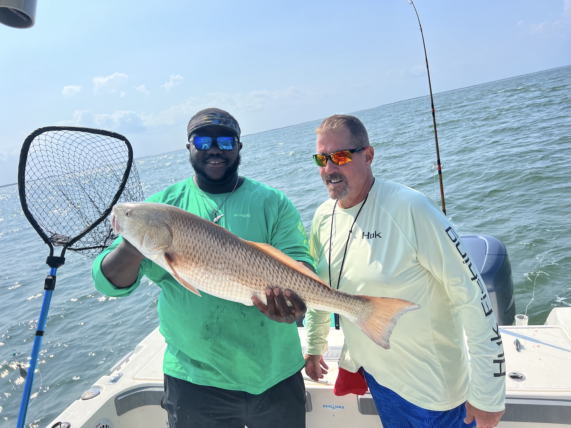 Two men on a boat at sea, one holding a large fish and the other holding a fishing net. Both are smiling and wearing sunglasses and long-sleeve shirts. A fishing rod is visible in the background.