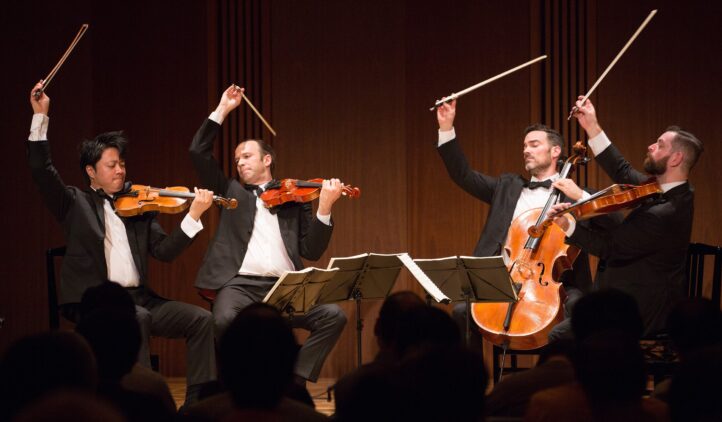 Four male musicians in tuxedos play string instruments on stage, raising their bows dramatically in unison, with sheet music in front of them and an audience in the foreground.