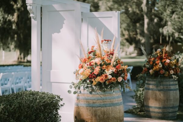 Two wooden barrels topped with vibrant floral arrangements stand beside an open white door frame outdoors. Rows of white chairs and lush greenery are visible in the background, suggesting a wedding or event setting.