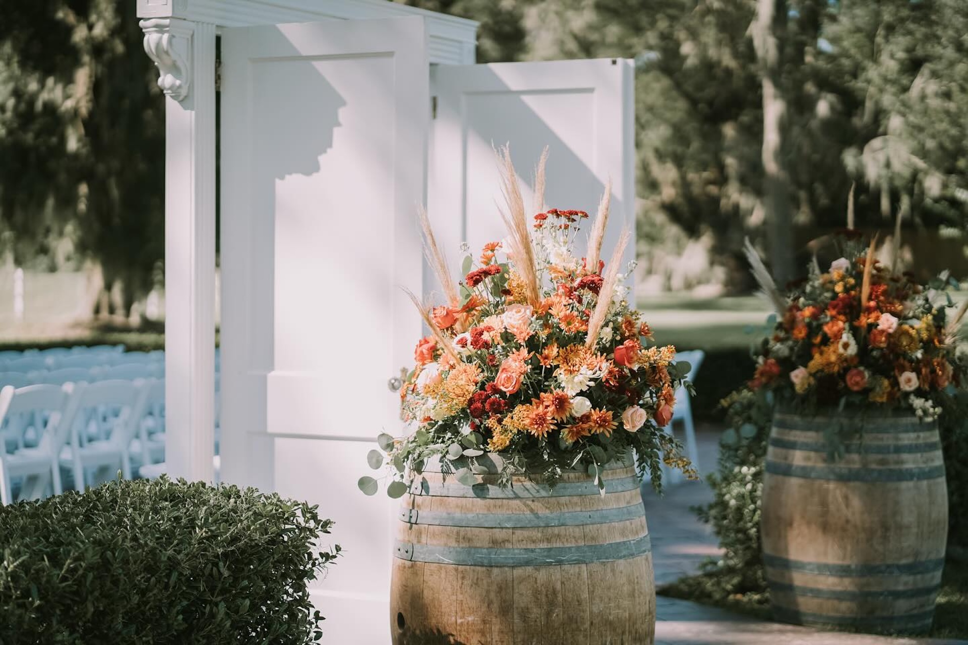 Two wooden barrels topped with vibrant floral arrangements stand beside an open white door frame outdoors. Rows of white chairs and lush greenery are visible in the background, suggesting a wedding or event setting.