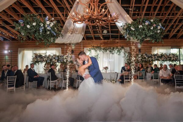 A bride and groom share their first dance in a rustic, decorated venue with wooden beams, floral arrangements, draped fabric, and artificial fog on the floor, surrounded by seated guests watching them.