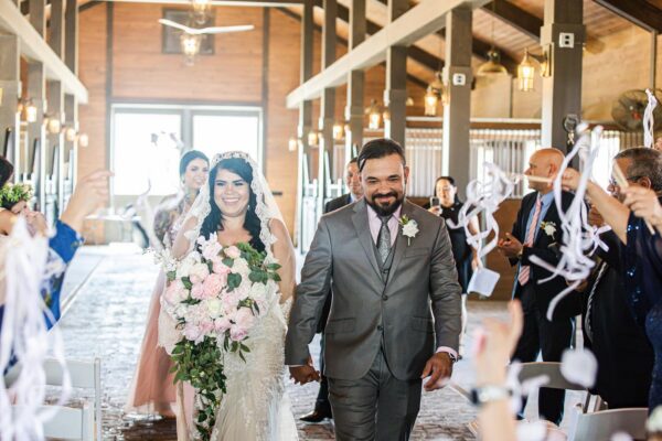 A bride and groom, smiling and holding hands, walk down the aisle after their wedding ceremony in a rustic venue as guests celebrate and toss white streamers. The bride holds a large bouquet of pink and white flowers.