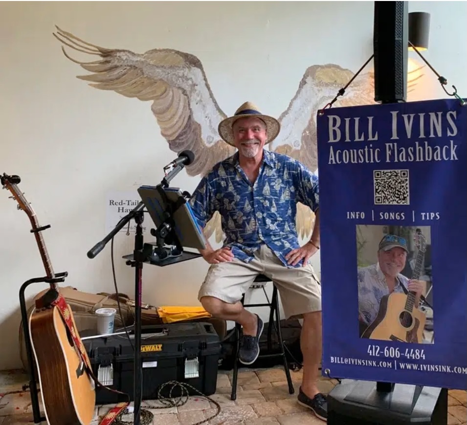 A smiling man in a blue patterned shirt and straw hat sits on a stool with musical equipment and a guitar nearby. A banner for Bill Ivins Acoustic Flashback stands in the foreground. A mural of bird wings is on the wall behind him.