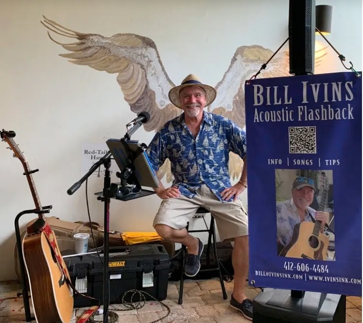 A smiling man in a straw hat sits with a guitar on his lap in front of a mural of outstretched bird wings, next to a sign reading Bill Ivins Acoustic Flashback with contact info and a photo of him playing guitar.