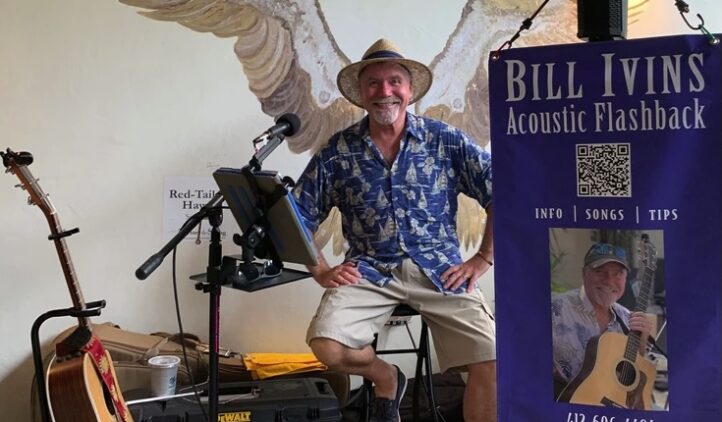 A smiling man in a blue shirt and straw hat sits in front of a microphone and music setup, with a guitar beside him and painted bird wings on the wall behind. A banner for Bill Ivins Acoustic Flashback is displayed on the right.