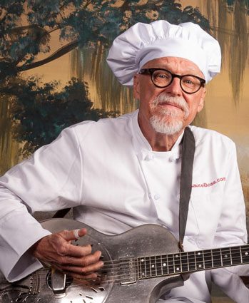 An older man in a chef’s hat and white coat, wearing glasses, sits holding a metal guitar. A soft-focus backdrop with trees and sky is behind him.