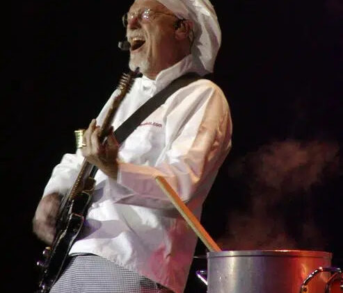 A musician dressed as a chef, wearing a white chef hat and coat, plays an electric guitar next to a steaming pot with a large wooden spoon on stage.