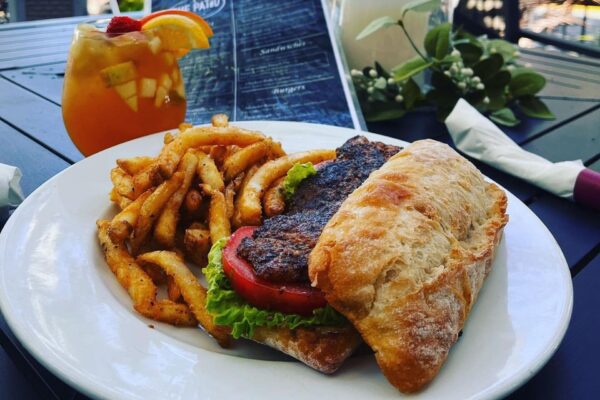 A plate with a toasted sandwich filled with lettuce, tomato, and a blackened patty, served with seasoned fries. A fruity cocktail and a menu are on the outdoor table in the background.