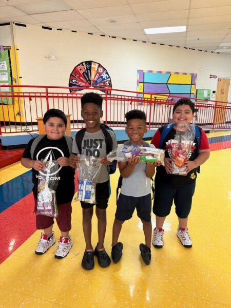 Four smiling boys stand indoors holding gift bags, posing together on a shiny, colorful floor near a red railing and a wheel decoration on the wall behind them.