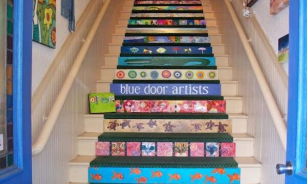 A staircase decorated with colorful, artistic designs on each step, including patterns of animals, fish, and abstract art. One step reads blue door artists in white text on a blue background.